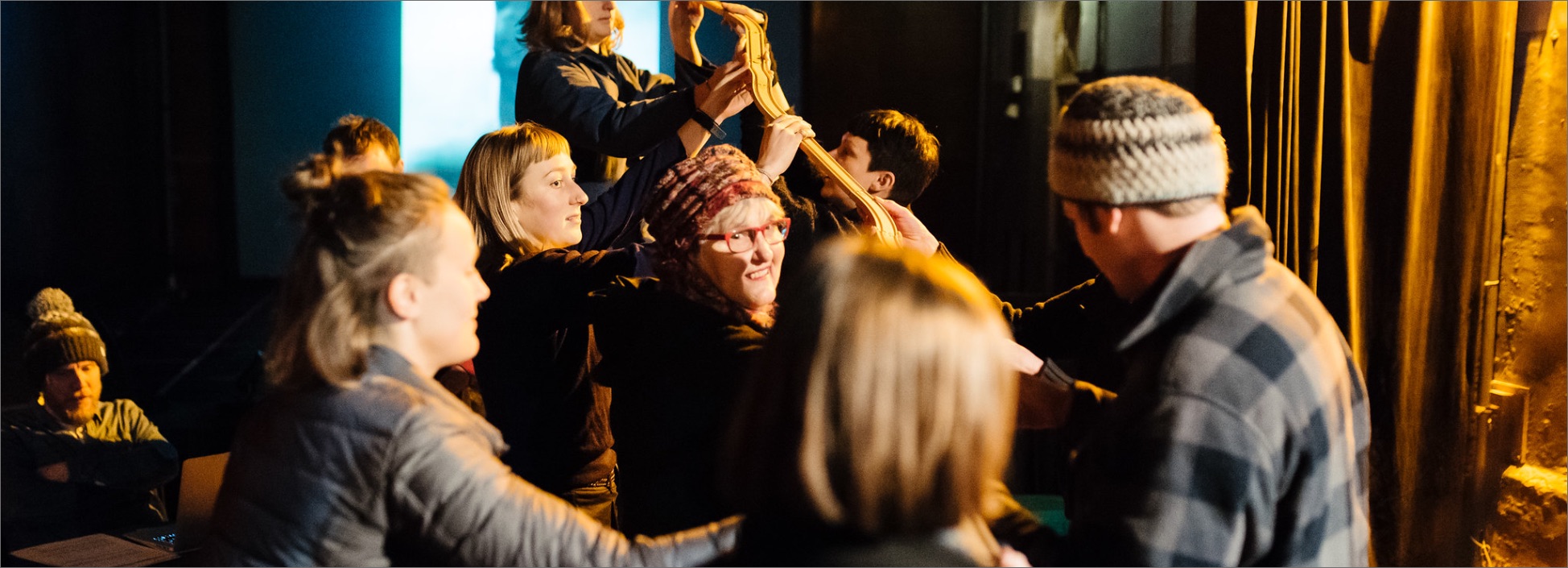 A group of adults of various ages hold wooden toy train tracks above their heads, trying to hold them together