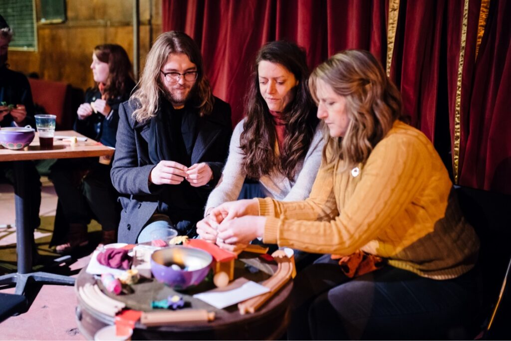 Three people sat at a table infront of a red velvet curtain. They are making something with plasterscine and toy train tracks