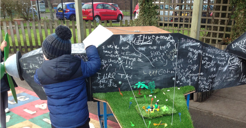A small boy faces a five foot long model of an aeroplane. The aeroplane is covered in chalk writing and has astro-turf on its wings with model people on