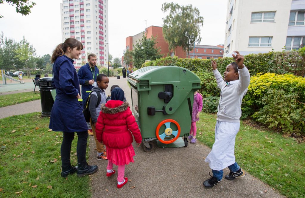 Five children and two adults stood on a path around a large green bin. One of the boys raises both arms in celebration