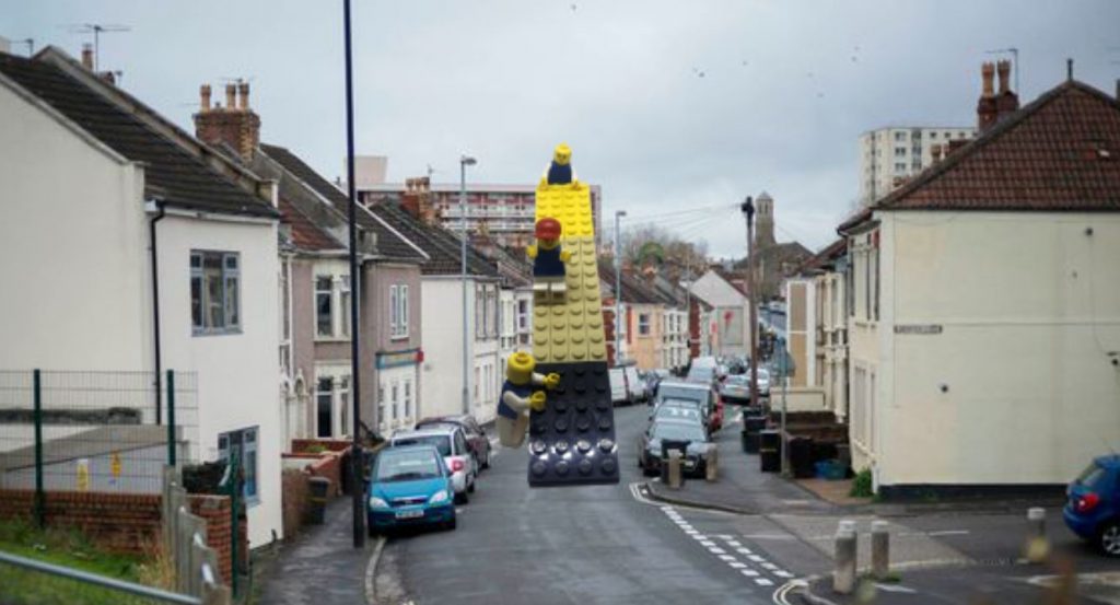 A street lined with terraced houses and parked cars. There is a photoshopped image of a slide made out of lego bricks in the middle of the road