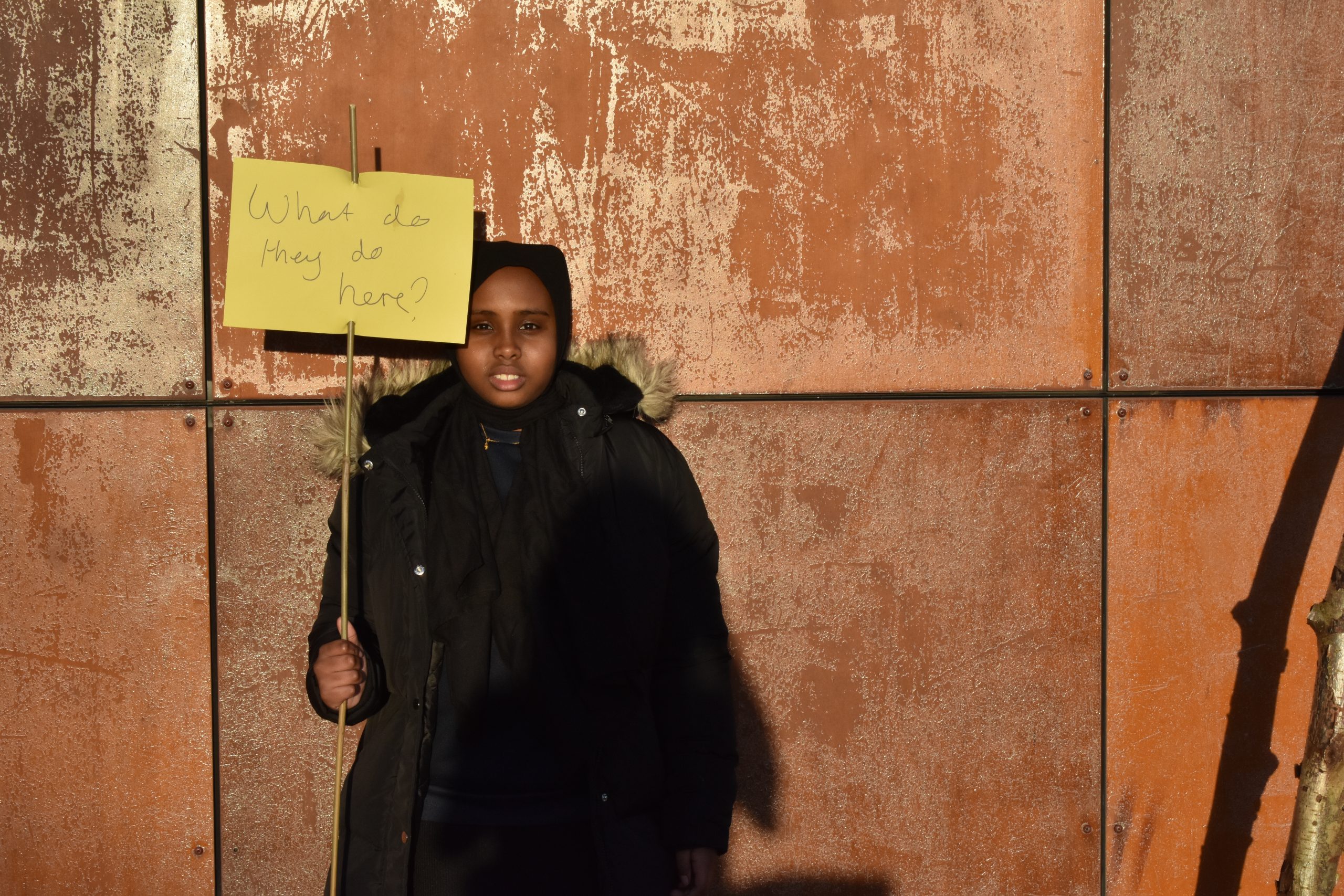 A child holds a yellow sign on a stick which says 'What do they do here?'
