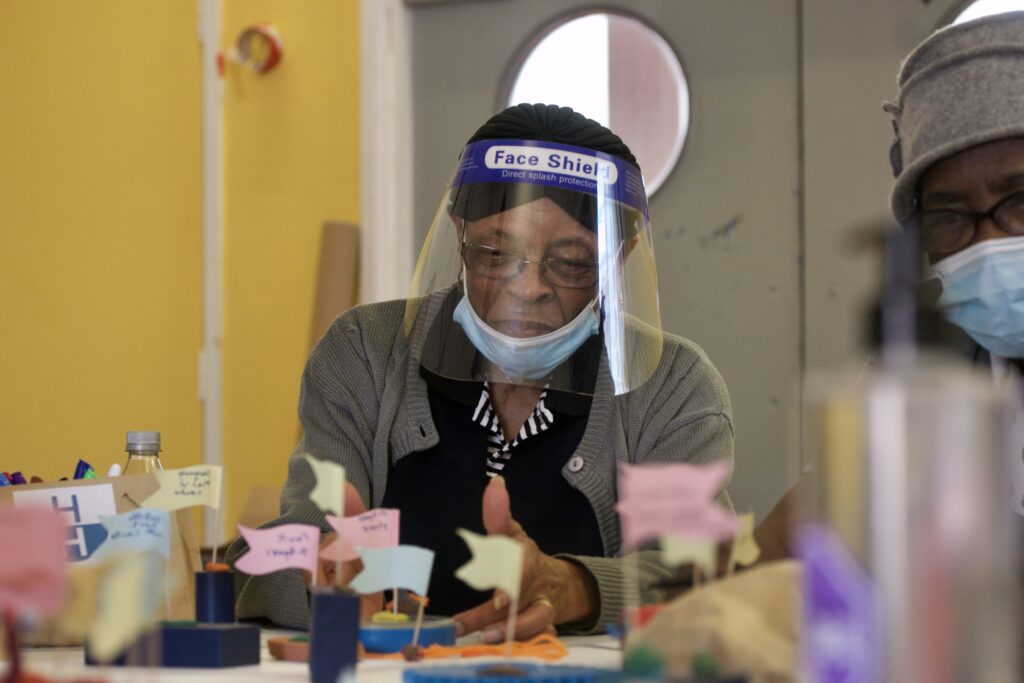 An older woman with a face visor gestures towards colourful block with colourful flags on the table infront of her