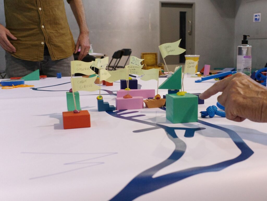 A hand points to colourful block and flags on a white table with a blue line representing the Avon River in Bristol on it