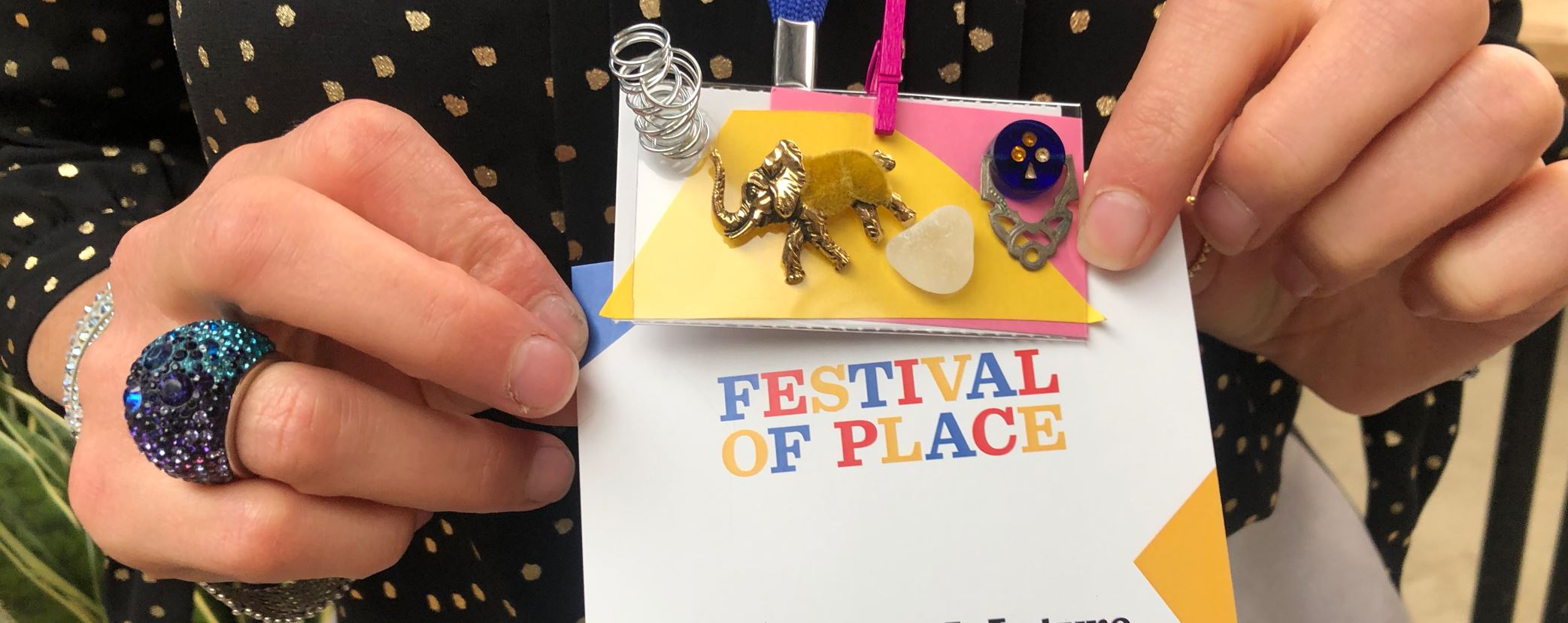 A small rectangular badge with objects stuck onto it including a small elephant, button and springs. Held up by hands infront of the words Fesitval of Place